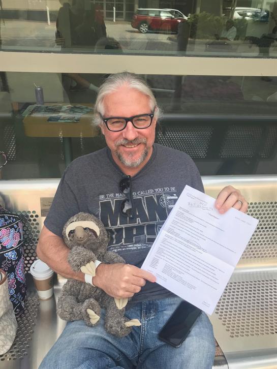 Smiling man displays a document while seated next to a person and holding a teddy bear outdoors.