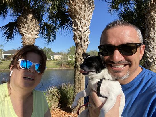 A couple by a sunny lake holds their small dog, with palm trees in the background.