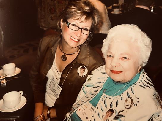 Two women smile together at a gathering, wearing name tags and dressed in stylish clothes.