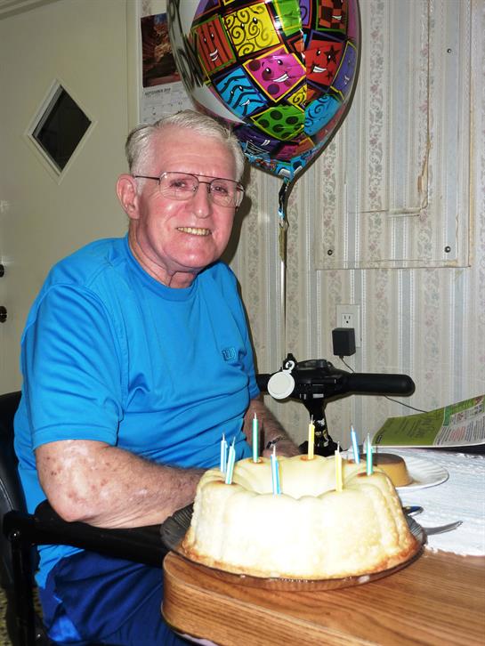 Smiling elderly man enjoys birthday cake with candles and colorful balloons in his home.