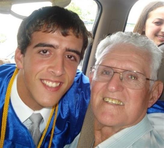 A graduate in blue robe smiles with a grandparent, celebrating in a car.