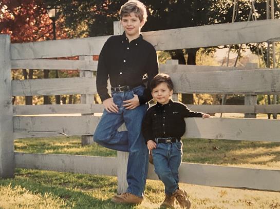 Boys dressed in matching outfits smile while posing near a fence in a colorful autumn setting.
