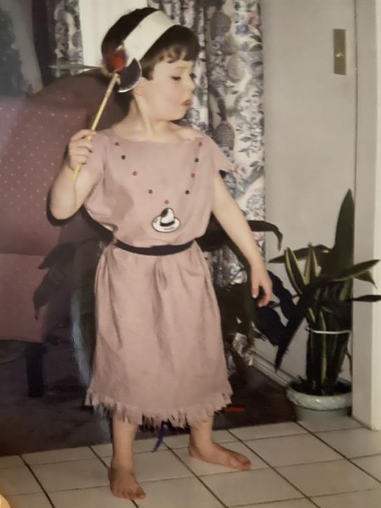 Young child in a playful outfit stands proudly with a wand, surrounded by indoor plants.
