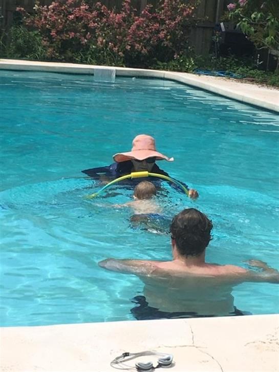 A child is learning to swim in a pool with help from an adult, enjoying the sunny weather.
