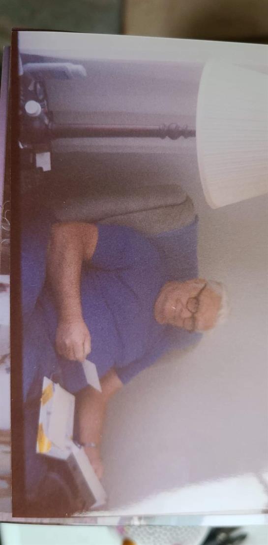 An elderly man with glasses cuts vegetables while seated at a wooden table in a warm room.