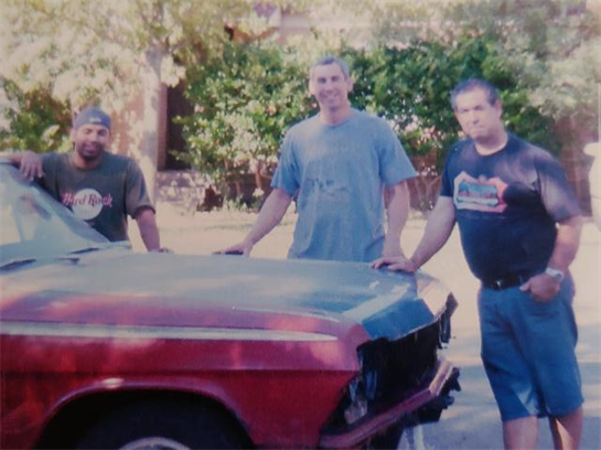 Three individuals gather around a classic car in a sunny outdoor setting, smiling and conversing.