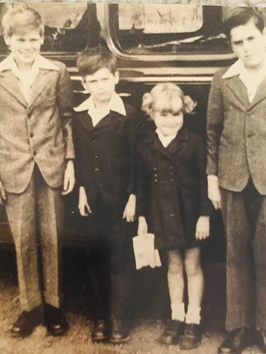 Four kids pose in vintage outfits by a classic car, capturing a nostalgic moment.