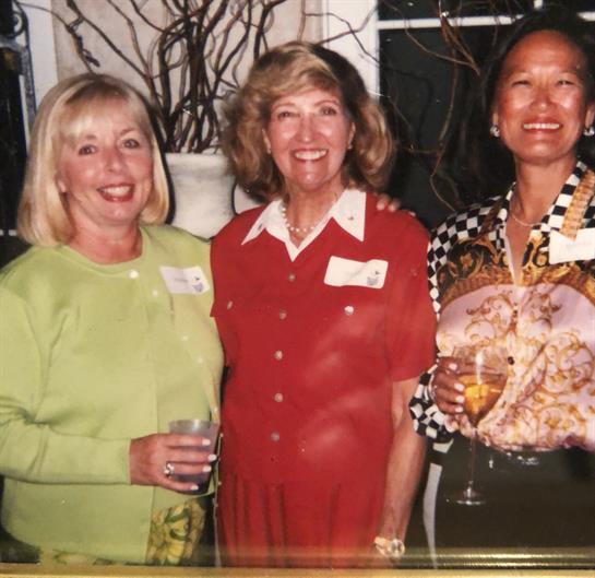 Three women smile together at a lively event, each holding a drink and dressed colorfully.