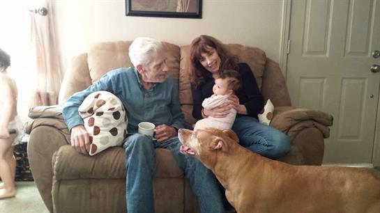 Elderly couple sits on a couch, smiling and interacting with a baby while a dog approaches.