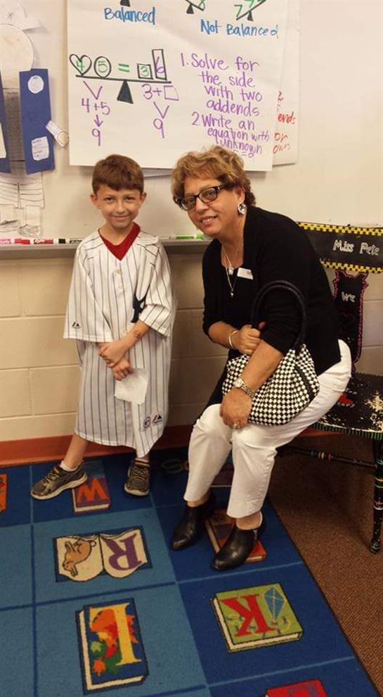 Young child in baseball attire stands next to a smiling woman in a classroom setting.