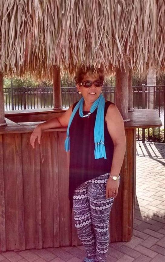 Brightly dressed woman stands at a wooden bar under a thatched roof by the water's edge.