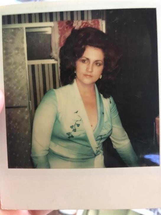 A woman in 1970s fashion sits elegantly, showcasing her intricate hairstyle in a dim room.