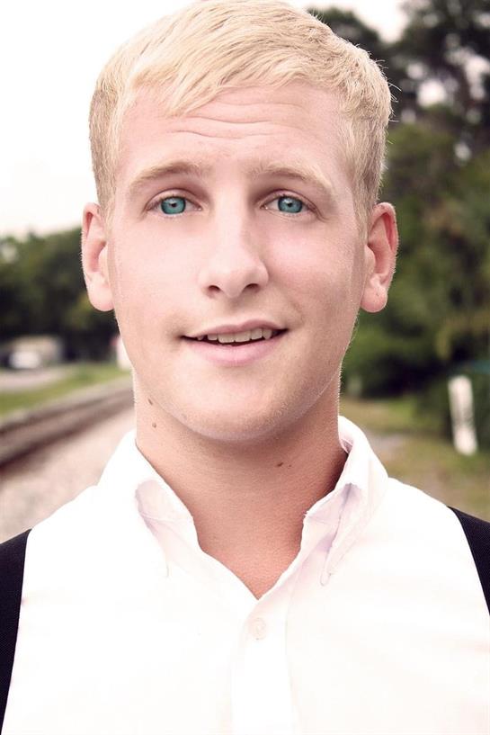 A young man with blue eyes stands confidently by railway tracks in bright daylight.