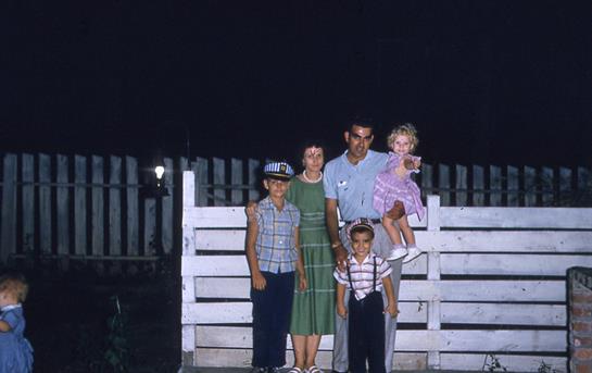 A family gathers outdoors at night, smiling and enjoying time together near a white fence.