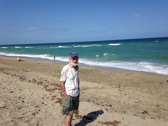 An elderly man stands on the sandy beach under a clear blue sky while waves crash nearby.