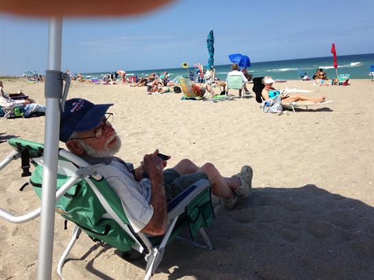 An elderly man sits comfortably in a chair at the beach, enjoying the sun and sea breeze.