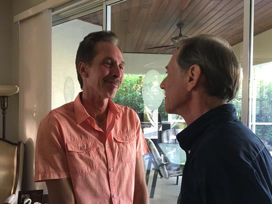 Two men engage in a warm conversation in a relaxed outdoor environment filled with plants.