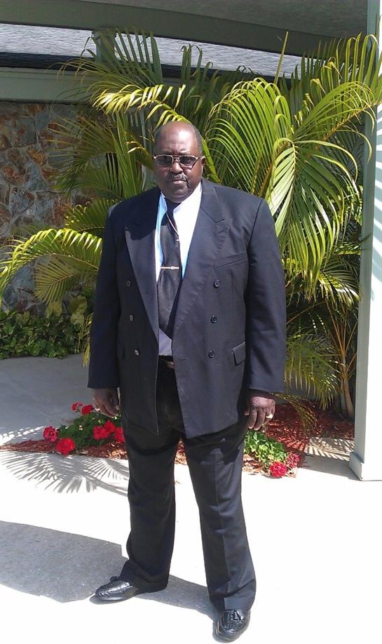 A well-dressed man poses confidently amidst vibrant greenery and blooming flowers.