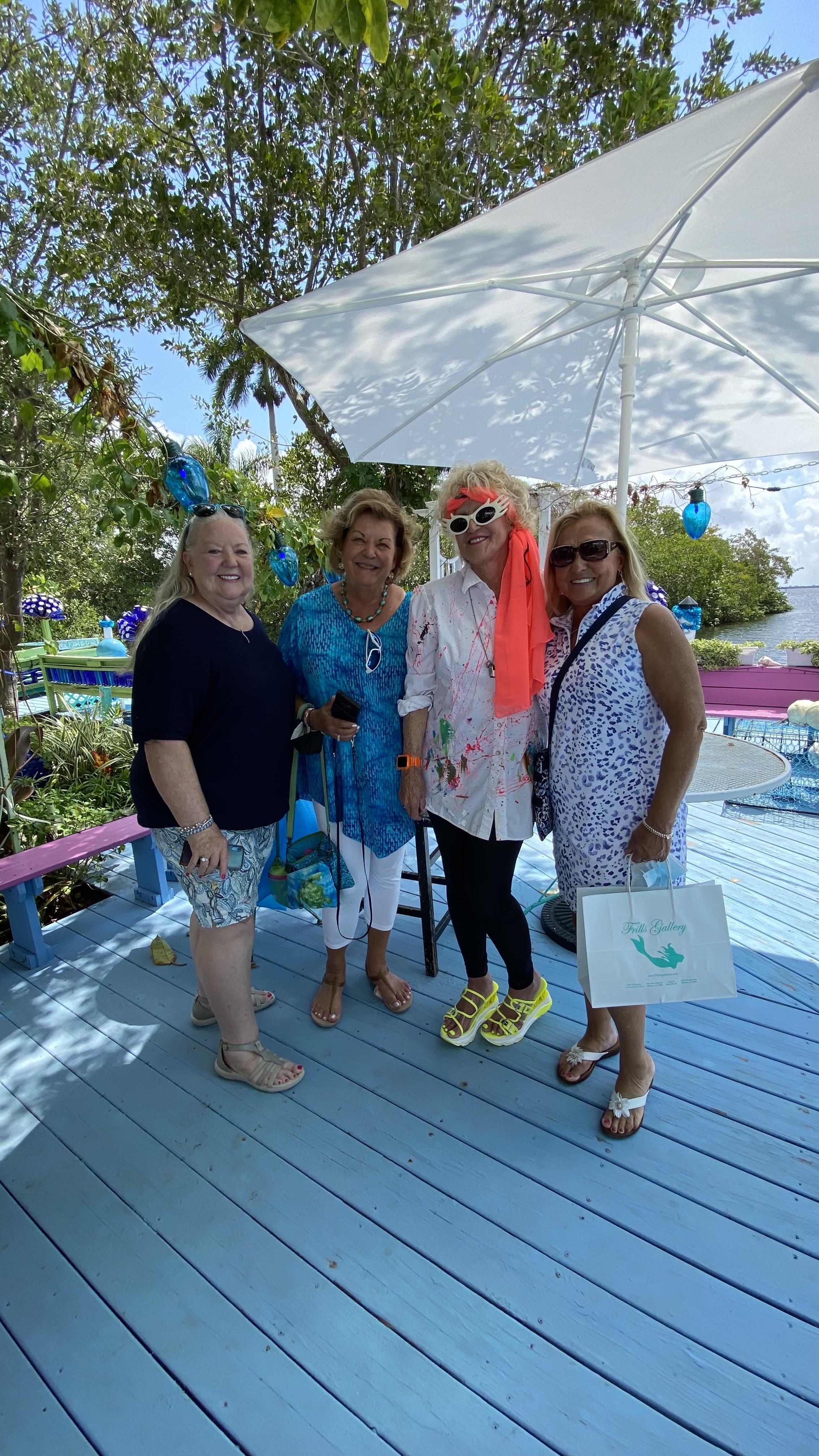 Four women smile and pose together on a bright deck under an umbrella, showcasing friendship.