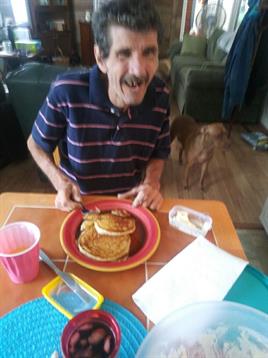 A man seated at a dining table enjoys pancakes, smiling and ready to eat in a cozy home.