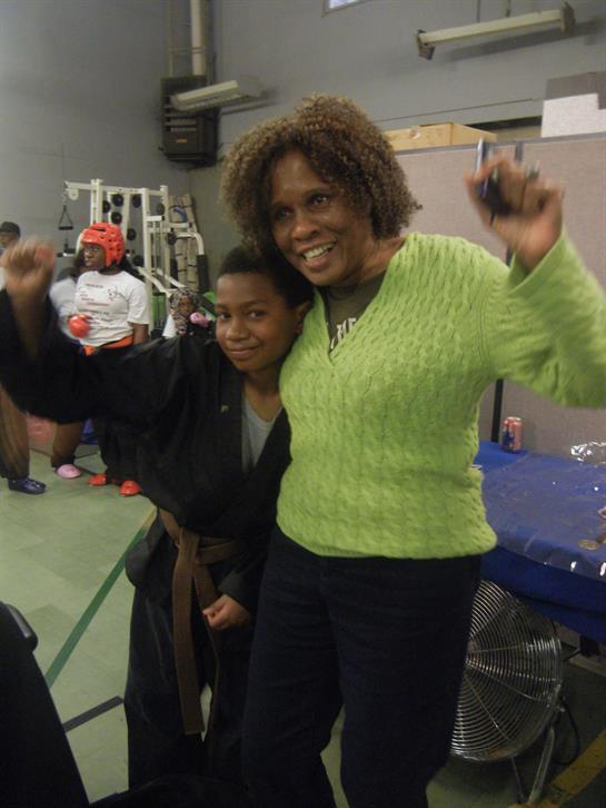 A young martial artist and mentor celebrate together in a gym, proudly raising their arms.