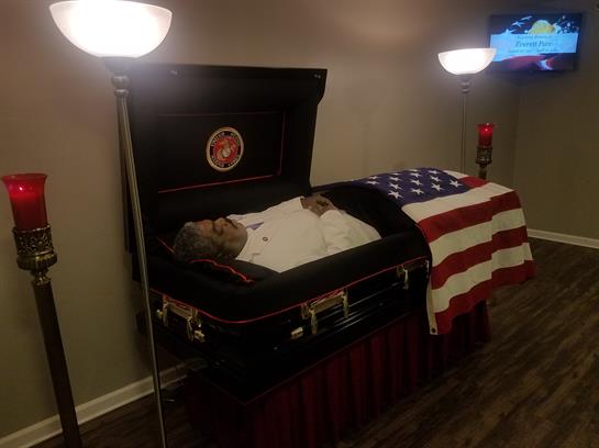 A veteran lies in a casket draped with an American flag, surrounded by lamps offering warm light.