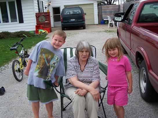 Two children pose happily next to an older woman sitting outdoors in a driveway.