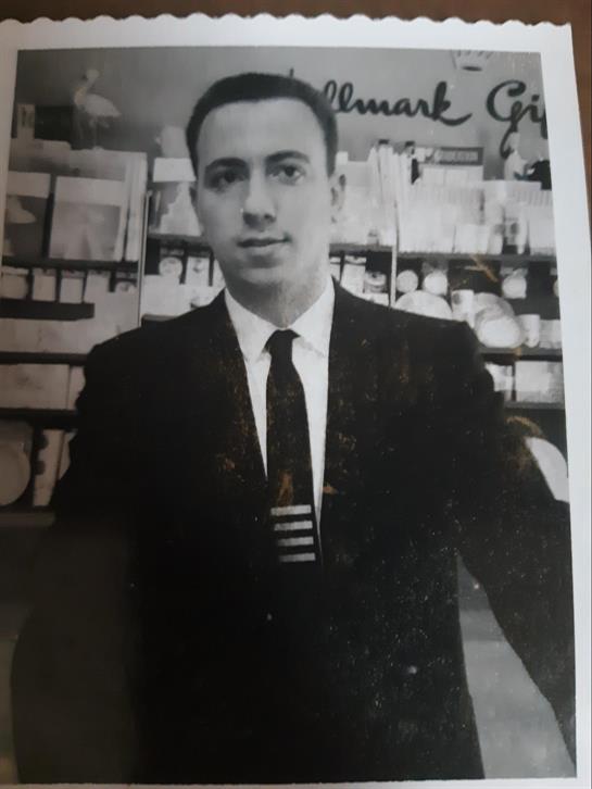 A young man poses confidently in formal clothing, surrounded by shelves filled with items.