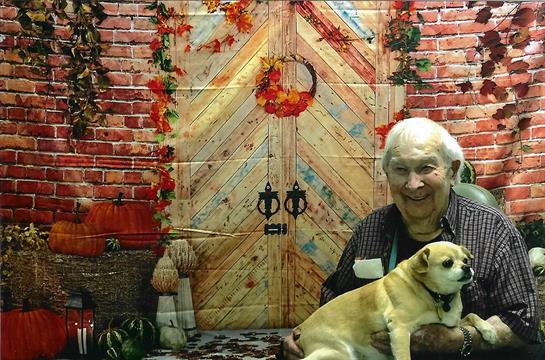 An elderly woman smiles while holding a small dog against a colorful autumn backdrop.