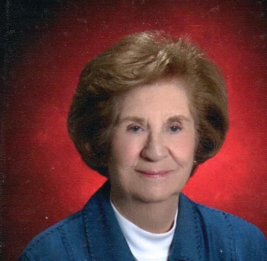 An elderly woman with brown hair smiles softly, wearing a blue jacket, set against a red backdrop.