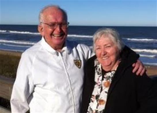 An elderly man and woman smile brightly while standing together by the shore on a sunny day.