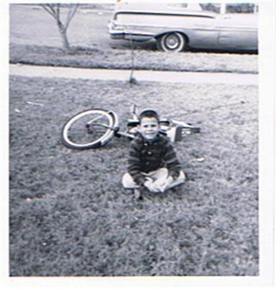 A boy smiles on the grass with a bicycle beside him and a classic car nearby.