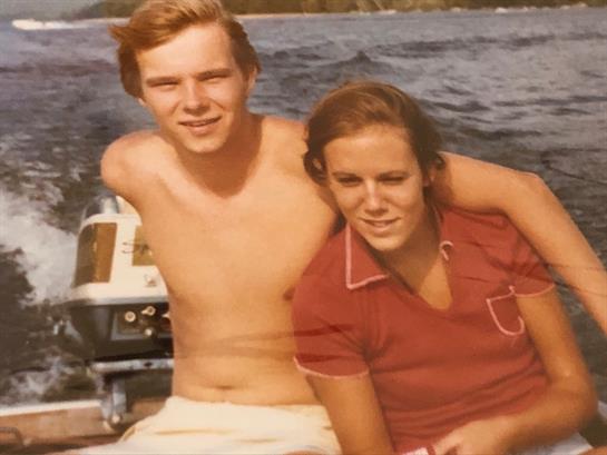 A young couple sits close together on a boat, smiling happily, surrounded by water and sunshine.