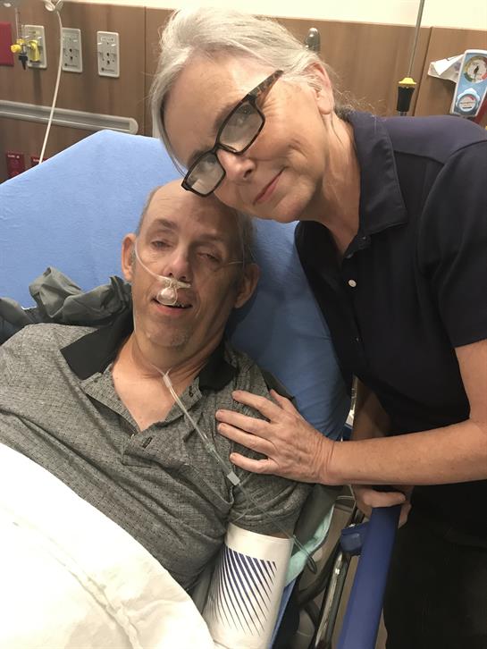 A caregiver smiles while offering comfort to a man in a hospital bed, showing compassion.