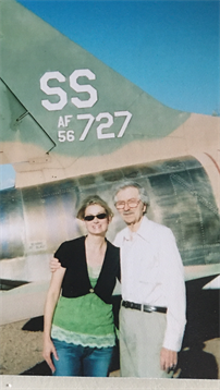 Couple stands together smiling in front of a military aircraft on a bright day.