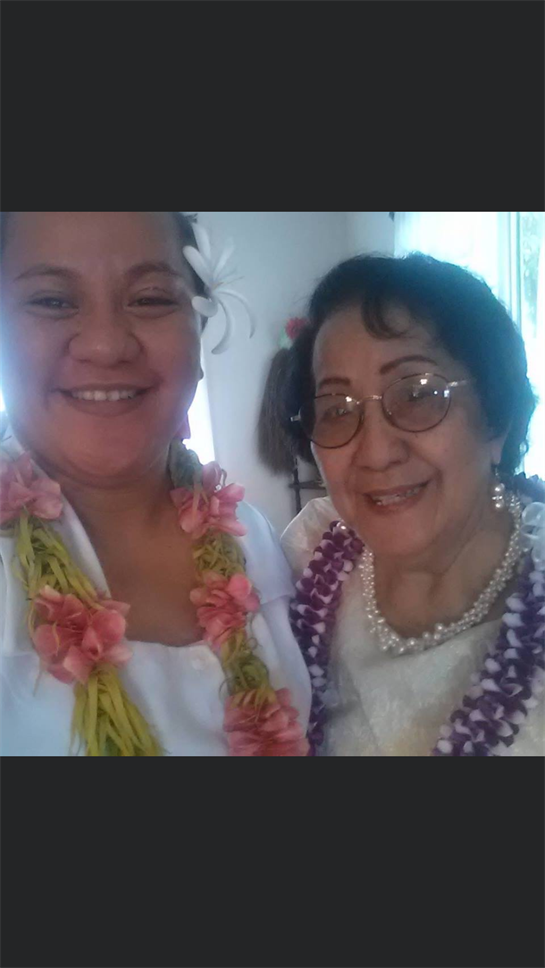 Two women embrace and smile at a joyful gathering, adorned in flower leis and bright attire.