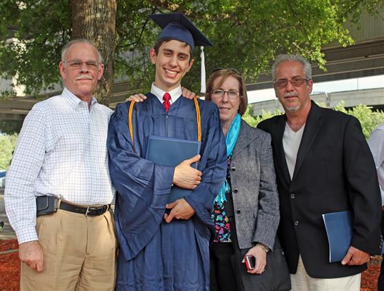 A young man in graduation attire stands with his family, smiling proudly in a park setting.