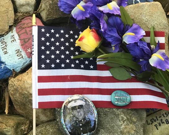 A small American flag stands among colorful flowers and stones, commemorating a fallen veteran.