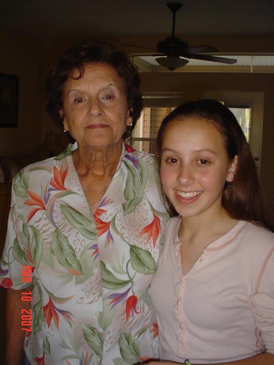 A senior woman stands beside a young girl, both smiling warmly in a welcoming living space.