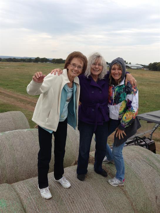 Three women share smiles and laughter while standing on hay bales in a field.
