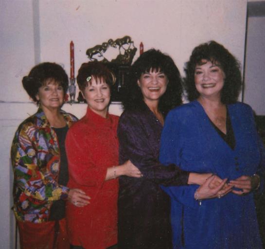 Four women stand closely together, dressed in colorful outfits, celebrating joyfully indoors.