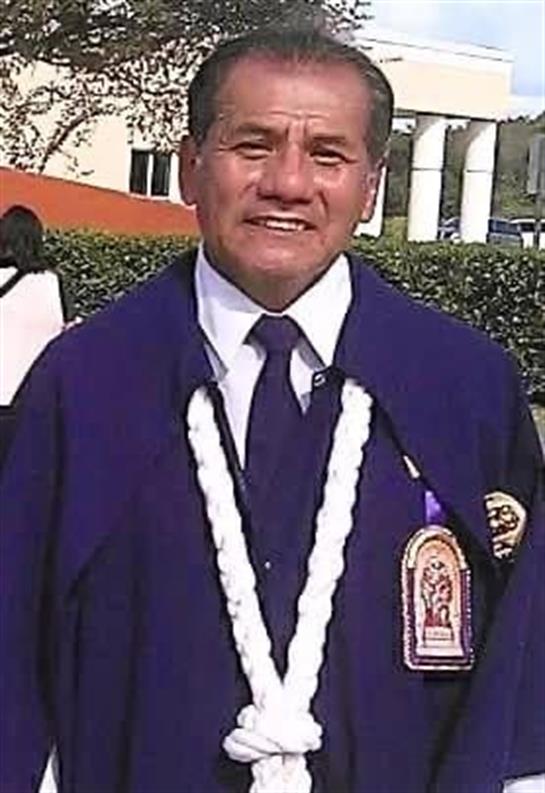 A man wearing traditional graduation robes and a ceremonial necklace smiles proudly on campus.