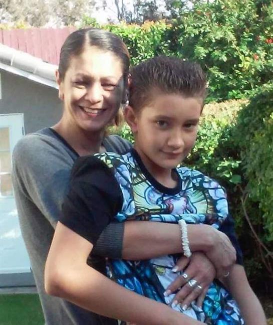 A smiling mother holds her son closely in a vibrant garden filled with greenery and sunlight.