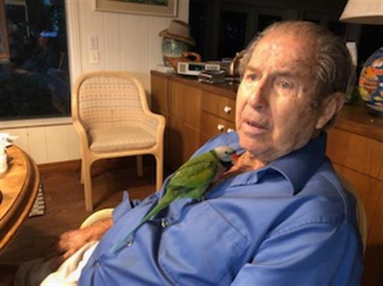 Elderly man relaxes in his home while a vibrant parrot perches playfully on his shoulder.