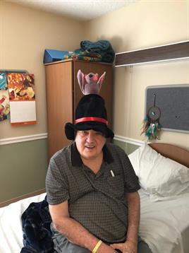 An older gentleman with a joyful expression is sitting on a bed, wearing a whimsical hat with a toy.