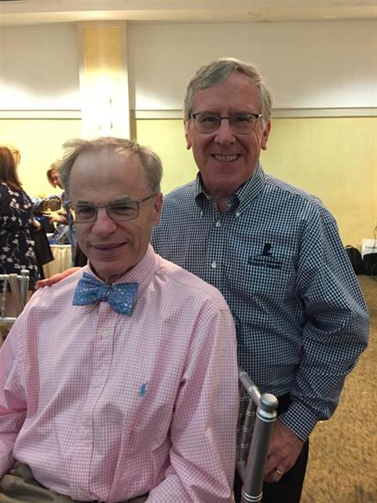 Two men stand together, both smiling joyfully, amidst a lively social gathering.