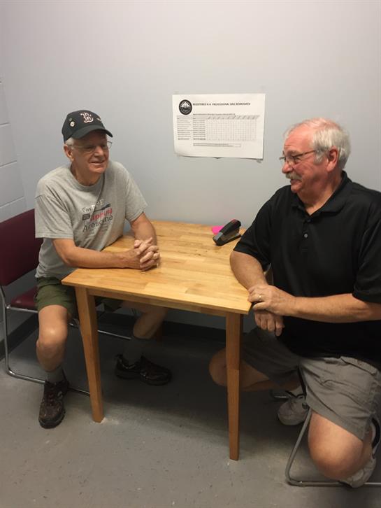 Two men chat amicably at a wooden table in a casual setting.