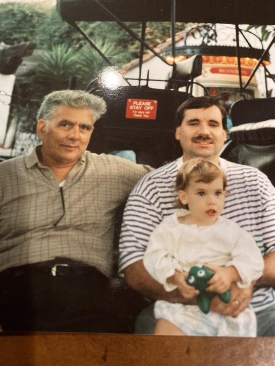 Two men seated beside a child in a park, smiling and enjoying a sunny day together.
