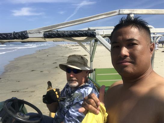 Two men are posing for a picture on a beach vehicle under the bright sun by the ocean.
