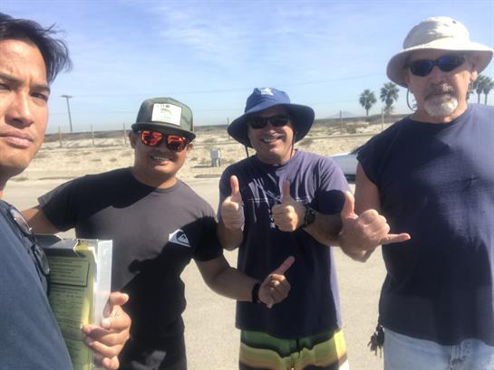 Four friends are smiling and giving thumbs up at the beach on a sunny day with palm trees.